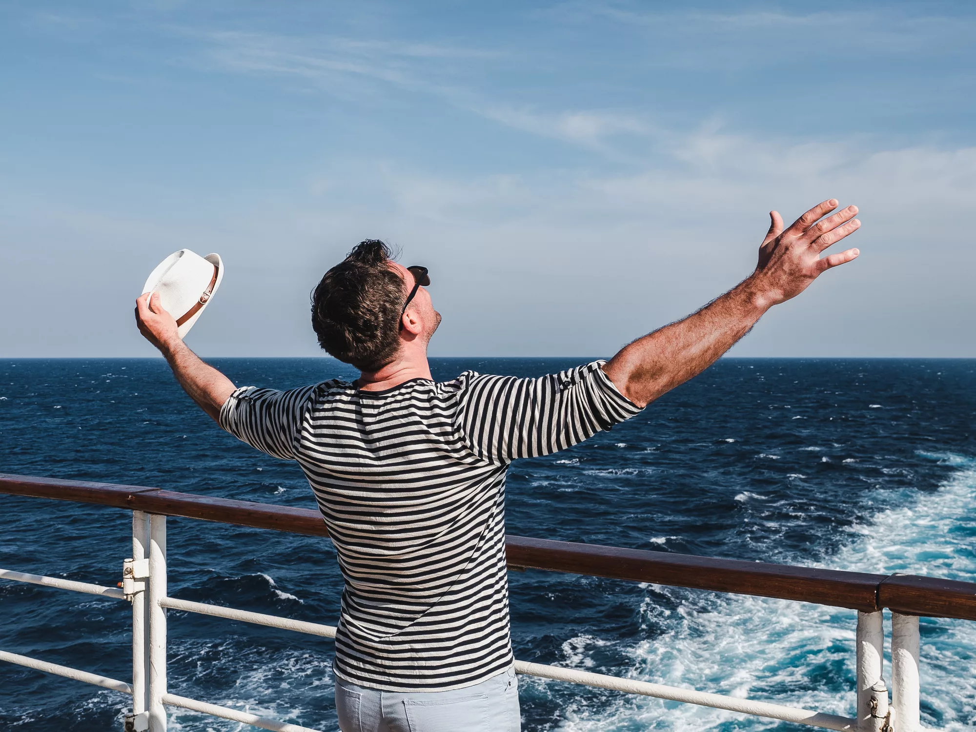 A male wearing a striped shirt looking out from the deck of a Mekong River cruise ship with his arms outstretched, holding a hat in one hand
